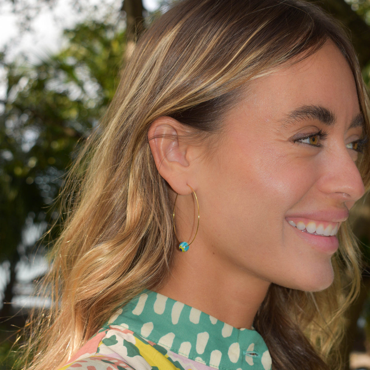 A woman in profile wearing a green and white patterned blouse and a delicate gold hoop earring with a small turquoise bead, photographed outdoors with trees in the background.