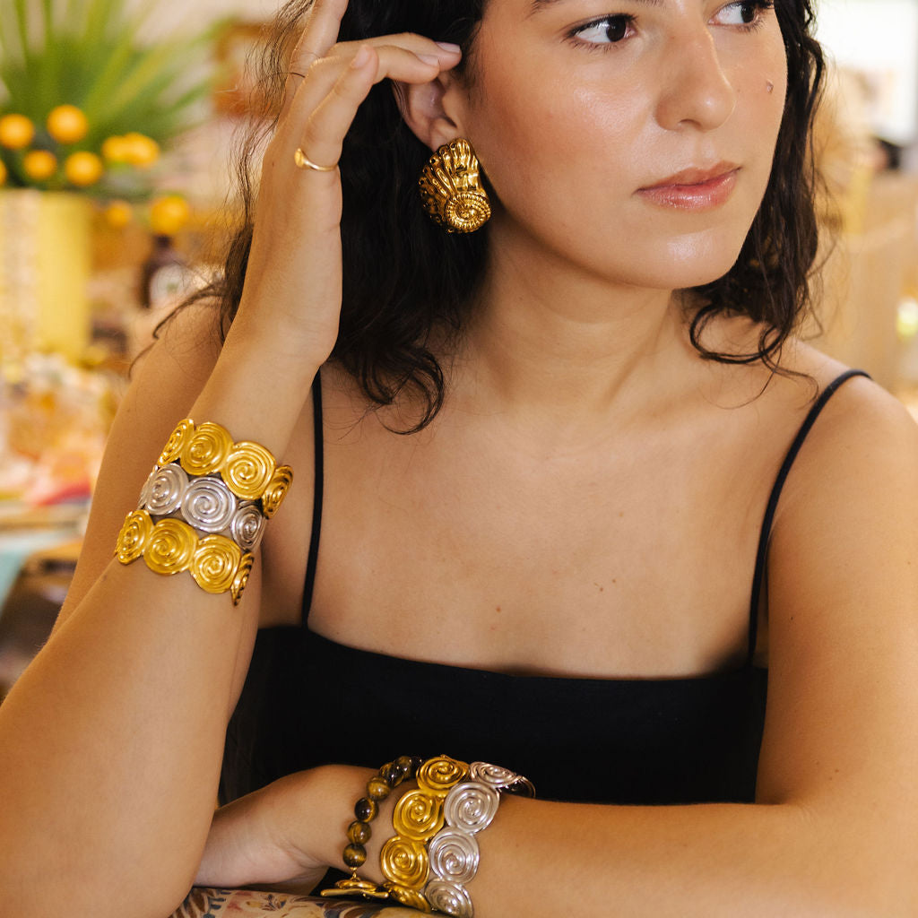 A woman wearing ornate gold and silver floral cuff bracelets and matching earrings, posing in a warm, softly blurred indoor setting.