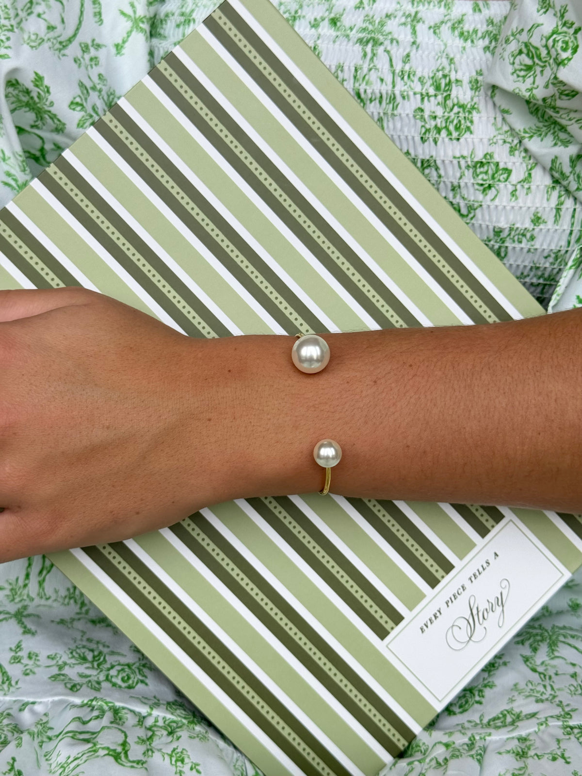 A person's forearm wearing delicate bracelet: The arm rests on a green and white striped book or box, with a green floral patterned fabric in the background.
