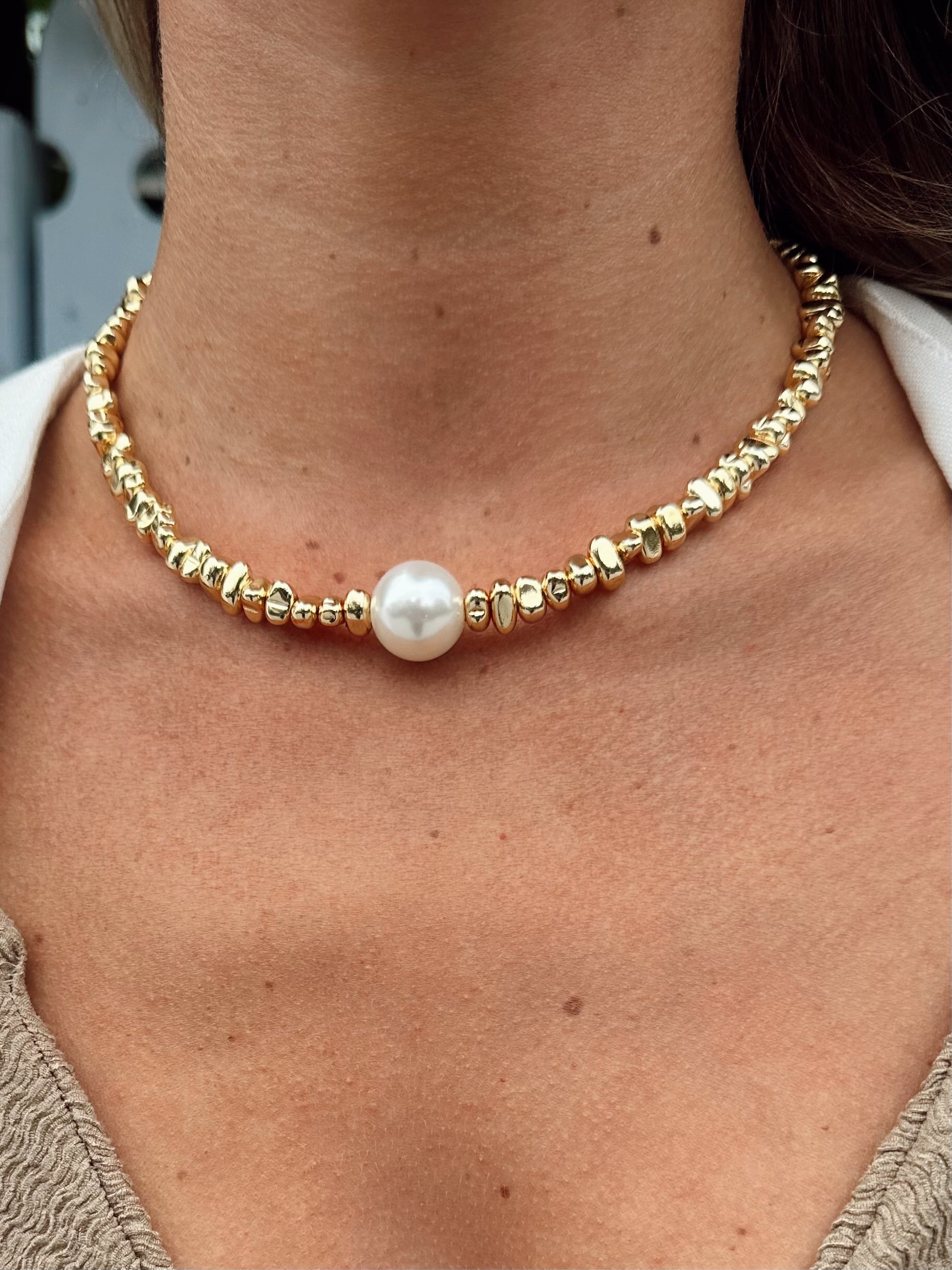 A close-up of a person's neck and upper chest wearing a gold beaded choker necklace with a single white pearl at the center.