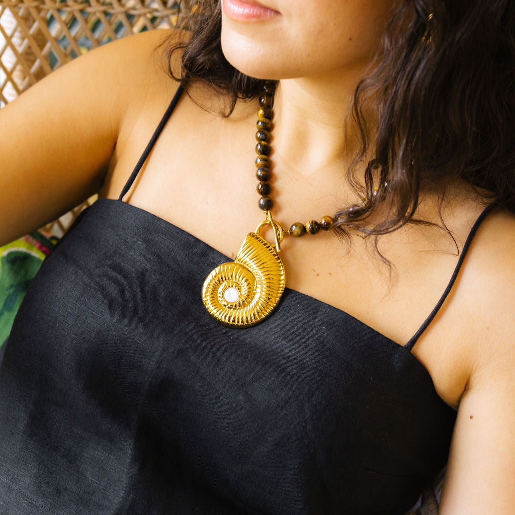 A close-up lifestyle shot of a woman wearing a black spaghetti-strap top, a brown beaded necklace with a large gold spiral pendant, and oversized clear-framed sunglasses.