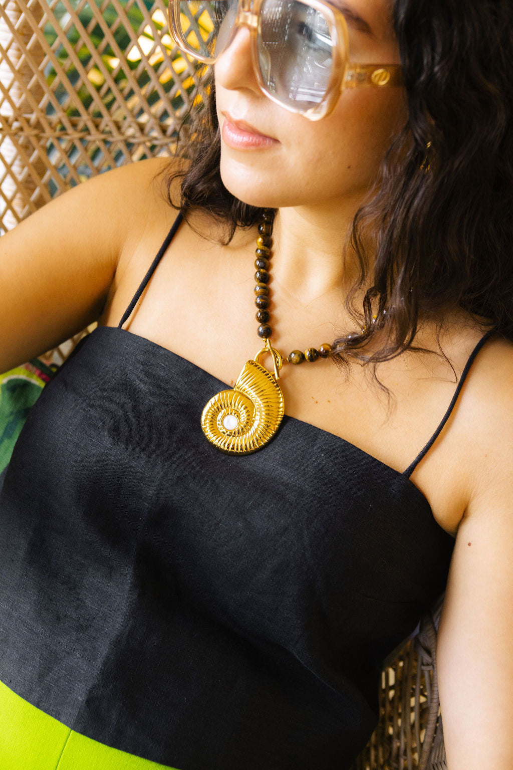 A close-up lifestyle shot of a woman wearing a black spaghetti-strap top, a brown beaded necklace with a large gold spiral pendant, and oversized clear-framed sunglasses.