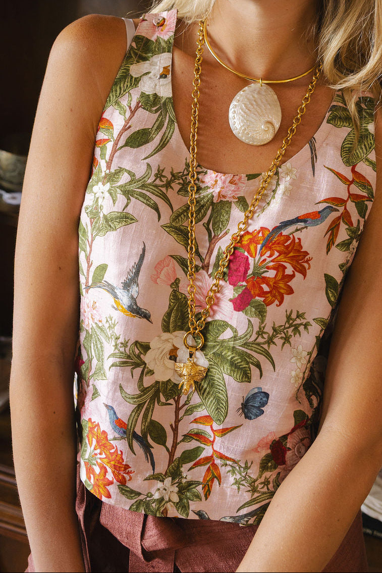 A woman wearing a sleeveless pink floral top with green leaves, orange flowers, and birds, accessorized with gold necklaces and sunglasses in a warm indoor setting.