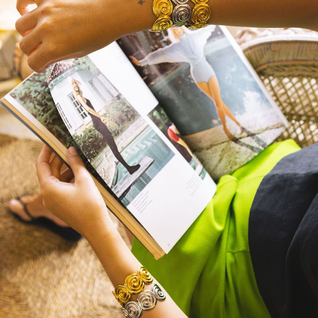 A close-up lifestyle shot of a person's arms and hands wearing two thick, decorative metallic bracelets while holding an open magazine. The bracelets feature a textured, layered design in gold and silver tones.