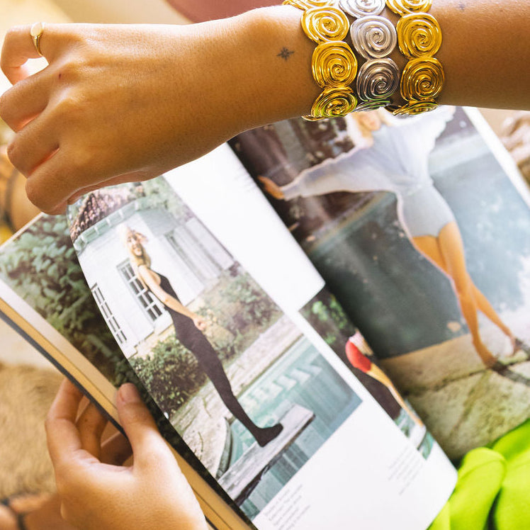 A close-up lifestyle shot of a person's arms and hands wearing two thick, decorative metallic bracelets while holding an open magazine. The bracelets feature a textured, layered design in gold and silver tones.