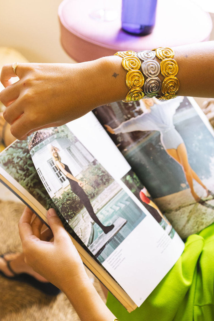 A close-up lifestyle shot of a person's arms and hands wearing two thick, decorative metallic bracelets while holding an open magazine. The bracelets feature a textured, layered design in gold and silver tones.