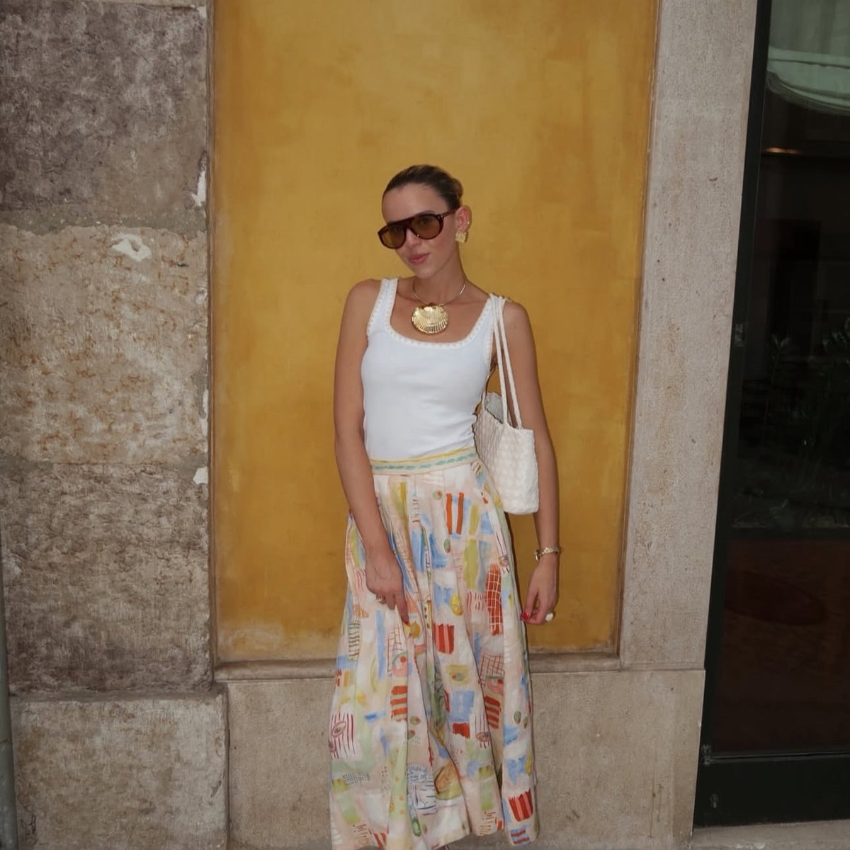Woman wearing a white top and colorful skirt against a yellow wall.