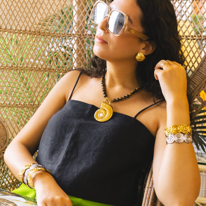 A woman wearing a black spaghetti-strap top and bright green skirt, sitting in a wicker hanging chair. She is wearing gold sunglasses, a gold spiral pendant necklace, and multiple gold and silver bracelets.