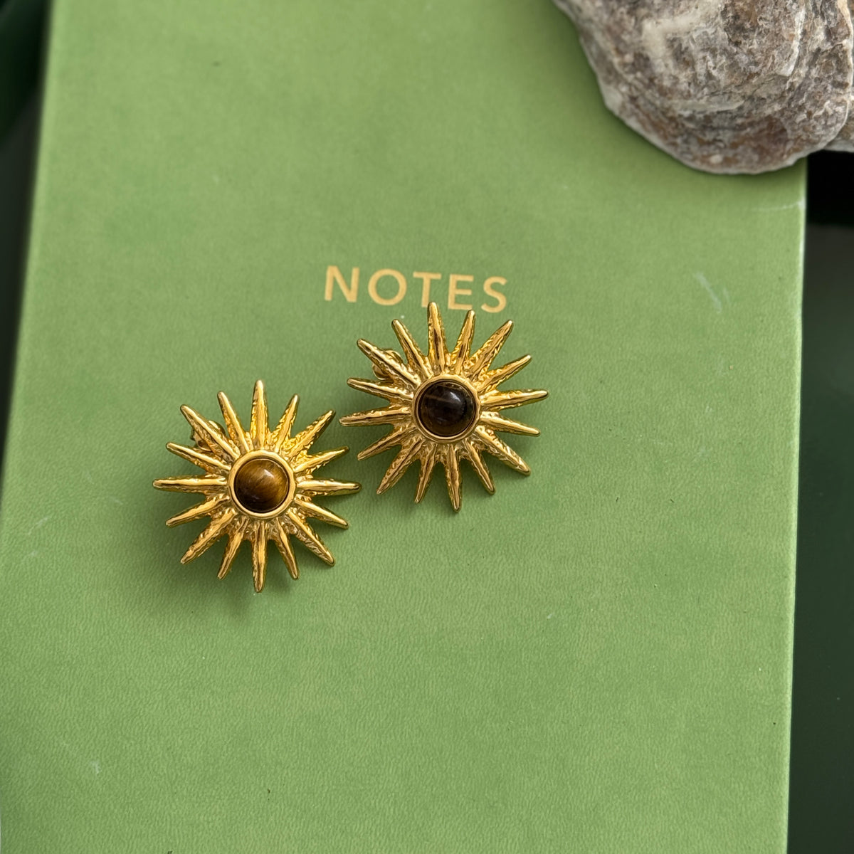 Gold sun-shaped earrings with a central brown gemstone on a green book.