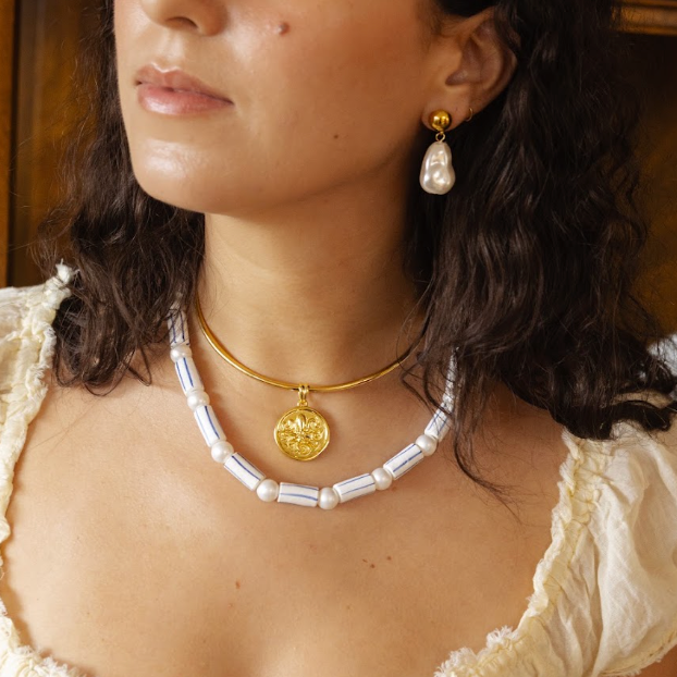 A woman wearing a gold necklace with a round medallion pendant and a white baroque pearl collar necklace, paired with a gold drop earring, in a warm indoor setting.