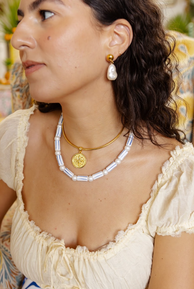 A close-up lifestyle shot of a woman wearing a gold necklace with a white baroque pearl pendant and a matching white baroque pearl drop earring, styled with a cream-colored ruffled blouse.