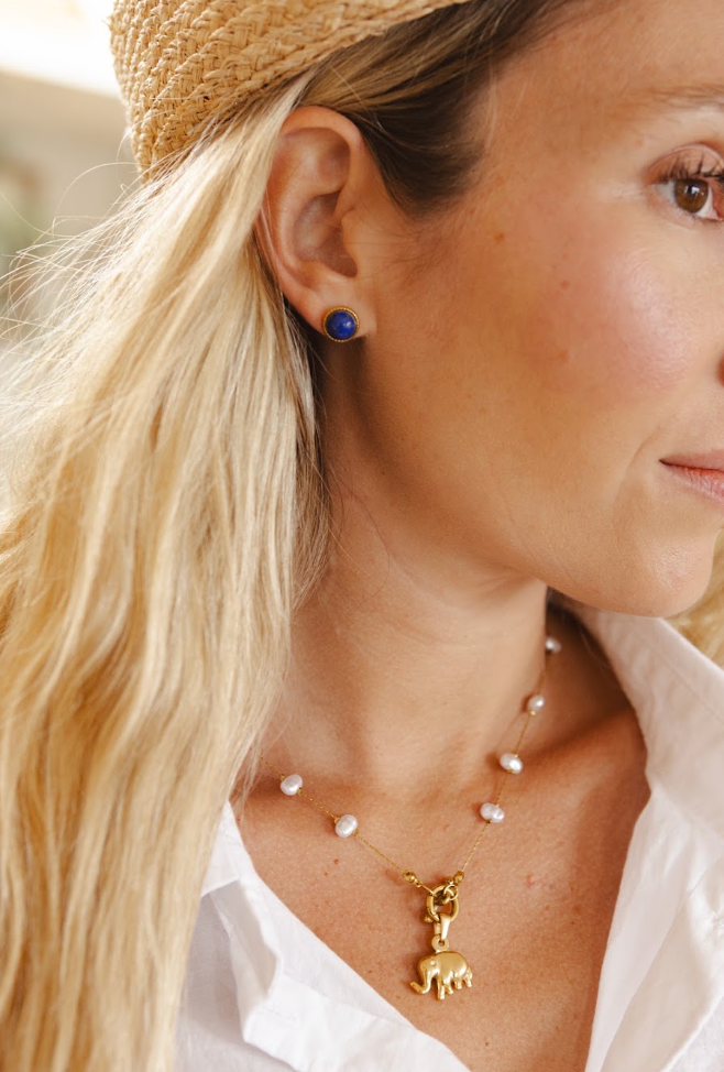 Close-up of a woman wearing a gold necklace with a small animal charm and white pearl beads, along with a straw hat and a blue stud earring.