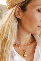 Close-up of a woman wearing a gold necklace with a small animal charm and white pearl beads, along with a straw hat and a blue stud earring.
