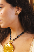 Close-up of a woman wearing a gold shell pendant necklace and matching gold stud earrings with dark beads.