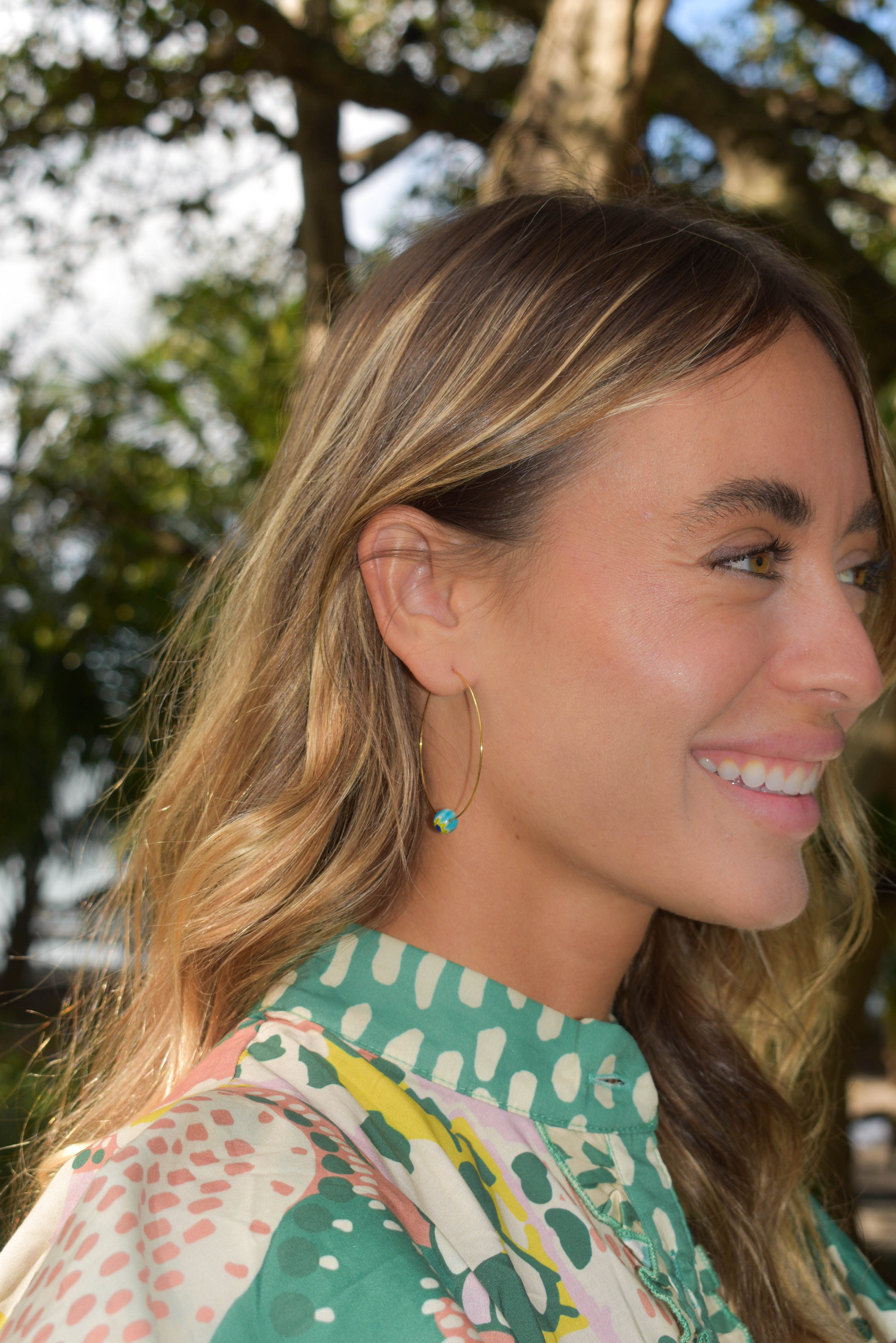 A woman in profile wearing a green and white patterned blouse and a delicate gold hoop earring with a small turquoise bead, photographed outdoors with trees in the background.