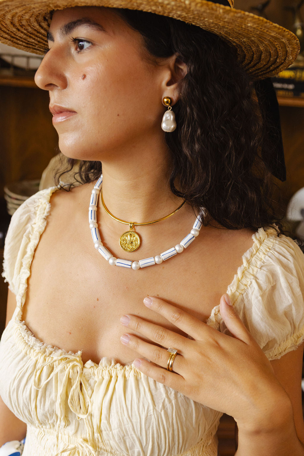 A woman wearing a straw hat, cream puff-sleeve top, and layered necklaces, including a white beaded collar necklace and gold medallion necklaces, along with matching gold and white earrings.