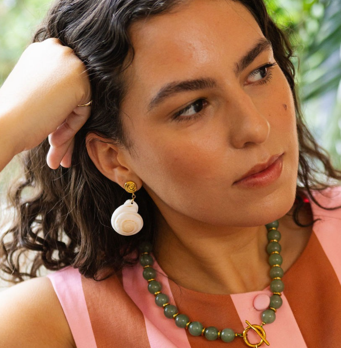 A close-up lifestyle shot of a woman wearing a green beaded necklace with gold accents and a matching drop earring, paired with a pink and brown striped top.