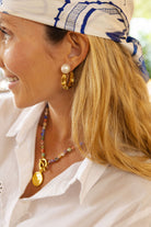 A close-up lifestyle shot of a woman wearing gold hoop earrings with white pearl studs, a beaded necklace with a gold medallion, and a white headscarf with blue patterns.