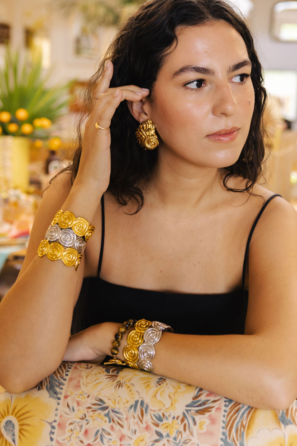 A woman wearing ornate gold and silver floral cuff bracelets and matching earrings, posing in a warm, softly blurred indoor setting.