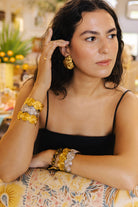 A woman wearing ornate gold and silver floral cuff bracelets and matching earrings, posing in a warm, softly blurred indoor setting.