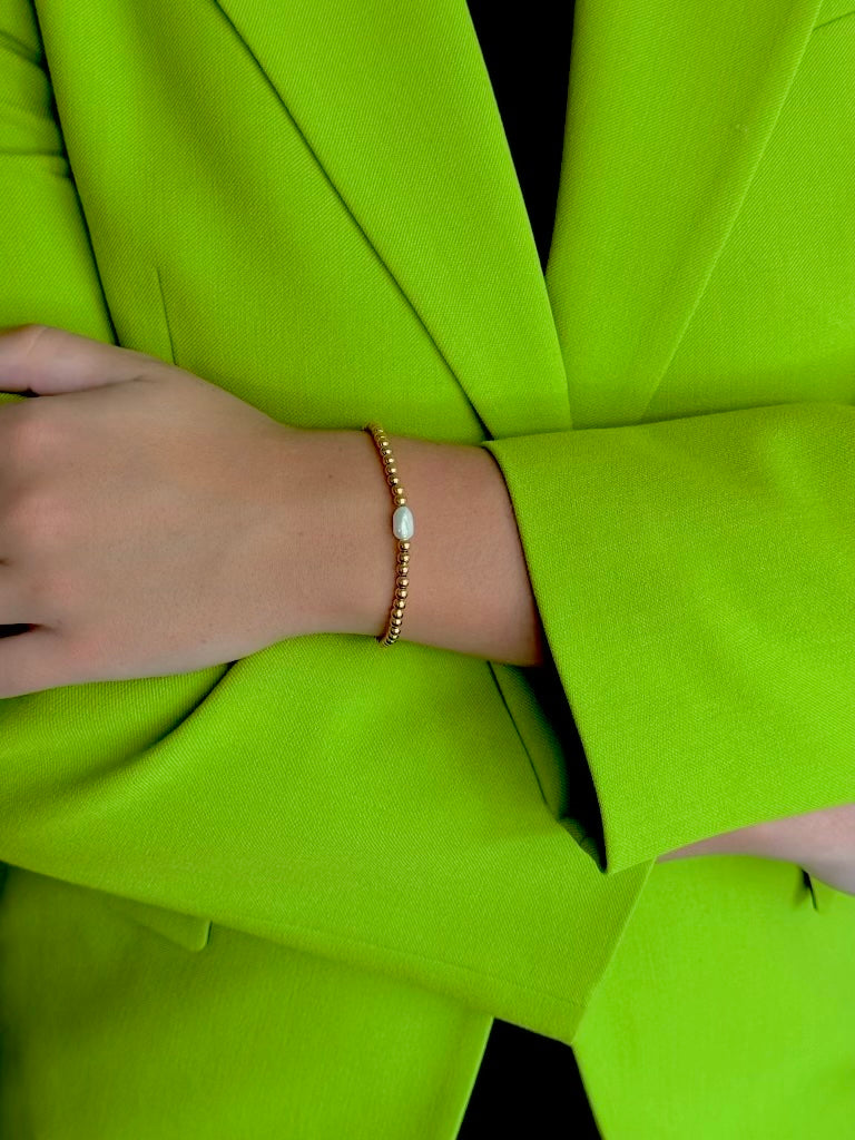 A close-up of a person's wrist wearing a delicate gold beaded bracelet with a small white oval stone, set against a bright lime green blazer.