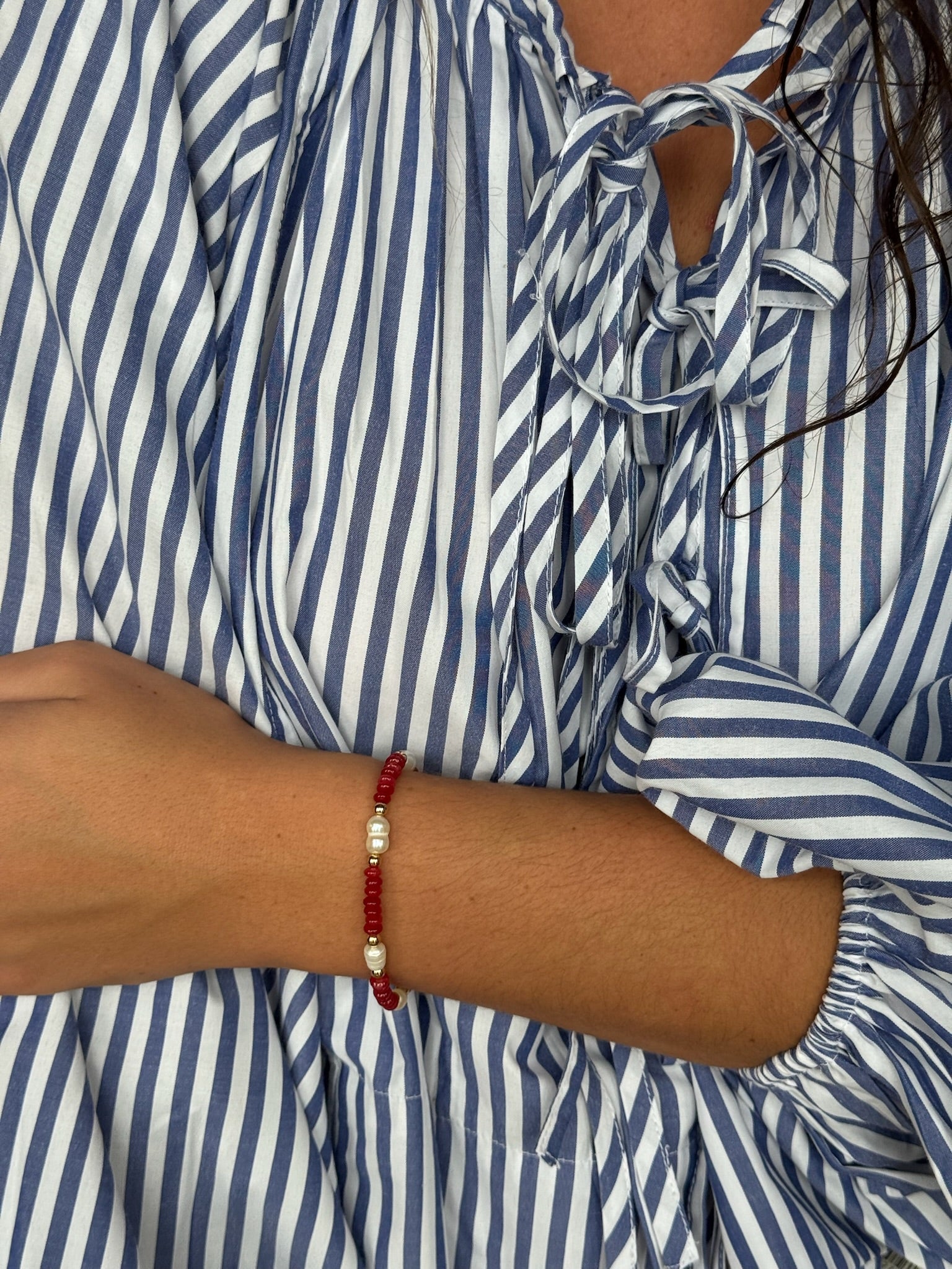 Lara Bracelet worn on the wrist – red beaded bracelet with pearl accents, styled with a blue and white striped blouse