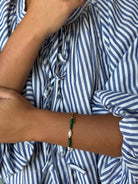A close-up of a person's wrist wearing a beaded bracelet with green beads, a white pearl, and a gold-toned ring, shown against a blue and white striped shirt.