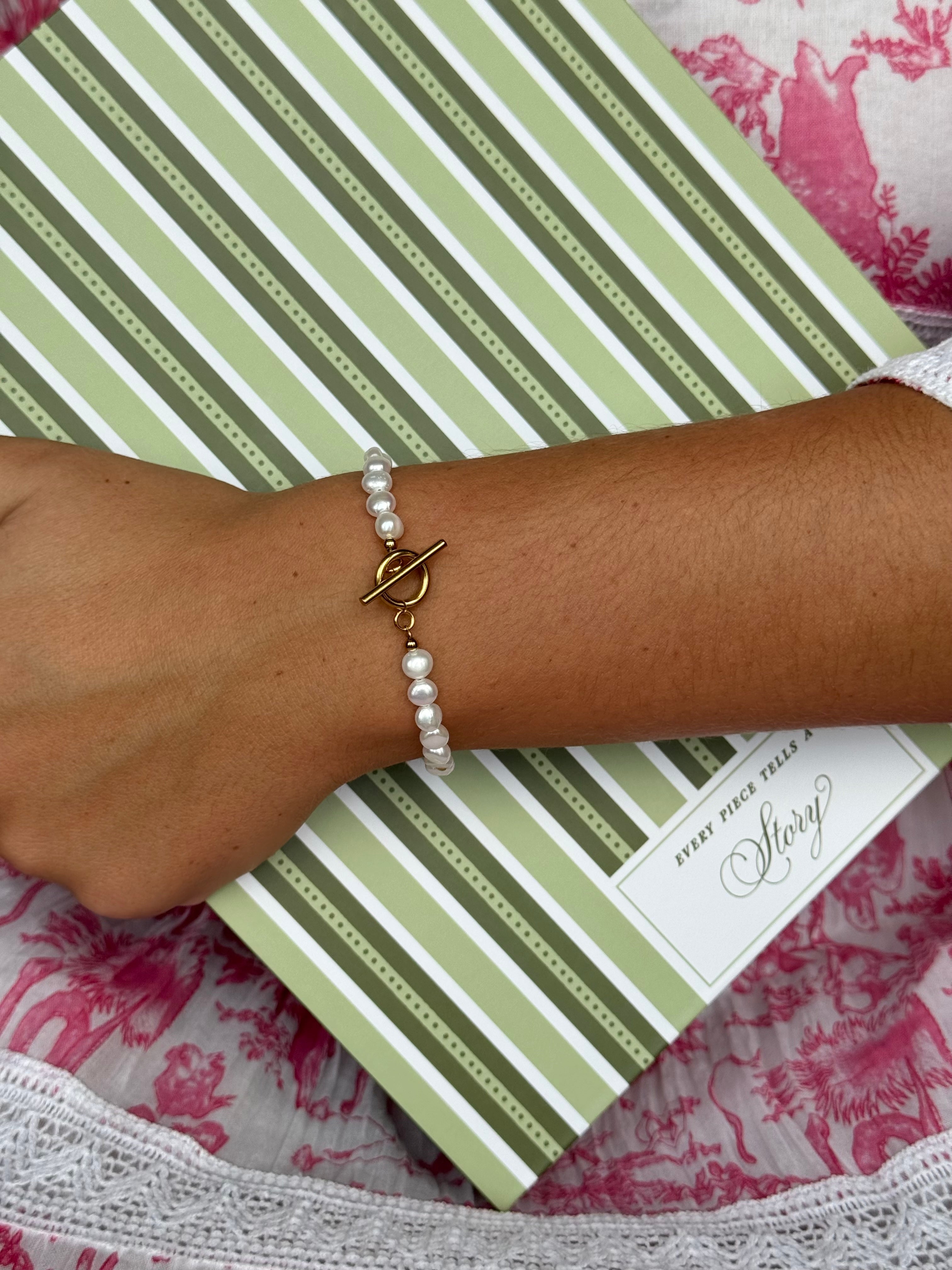 A close-up of a person's wrist wearing a pearl bracelet with a gold toggle clasp, resting on a green and white striped book or notebook over a pink and white patterned fabric.