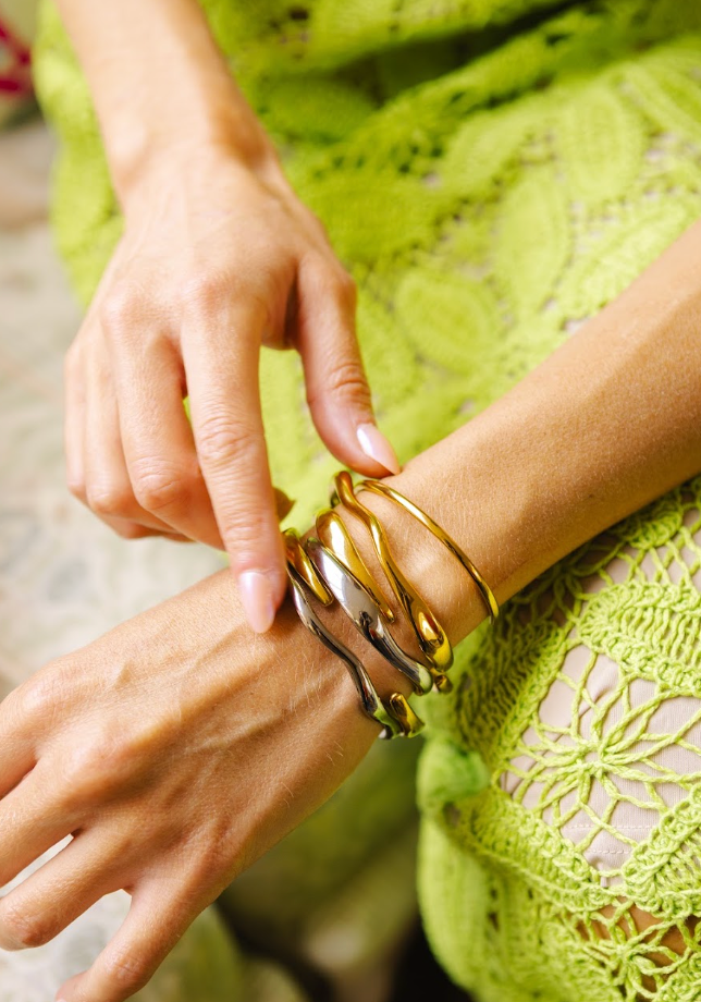 A close-up of a person wearing multiple metallic bangle bracelets on their wrist, with one hand touching the bracelets. The person is wearing a bright lime green crochet dress, and the background is a softly blurred neutral indoor setting.