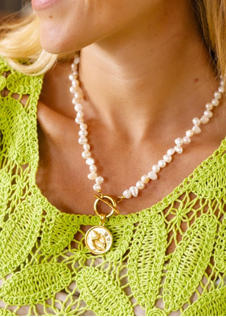 A close-up lifestyle shot of a woman wearing a white freshwater pearl necklace with a gold clasp and a gold pendant featuring a classical bust, paired with a matching pearl stud earring and a lime green crochet top.