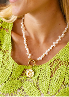 A close-up lifestyle shot of a woman wearing a white freshwater pearl necklace with a gold clasp and a gold pendant featuring a classical bust, paired with a matching pearl stud earring and a lime green crochet top.