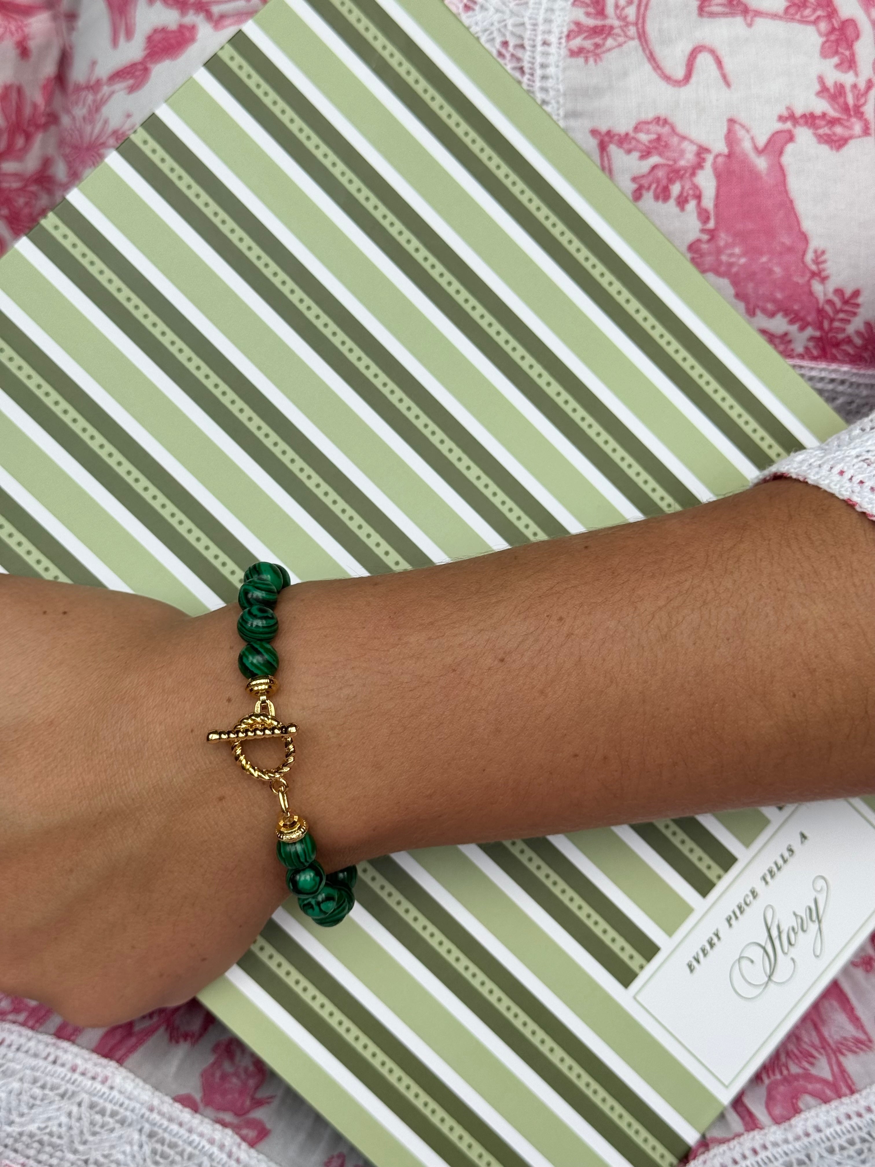 A close-up of a person's wrist wearing a green beaded bracelet with a gold toggle clasp, resting on a green and white striped book over a pink and white patterned textile.