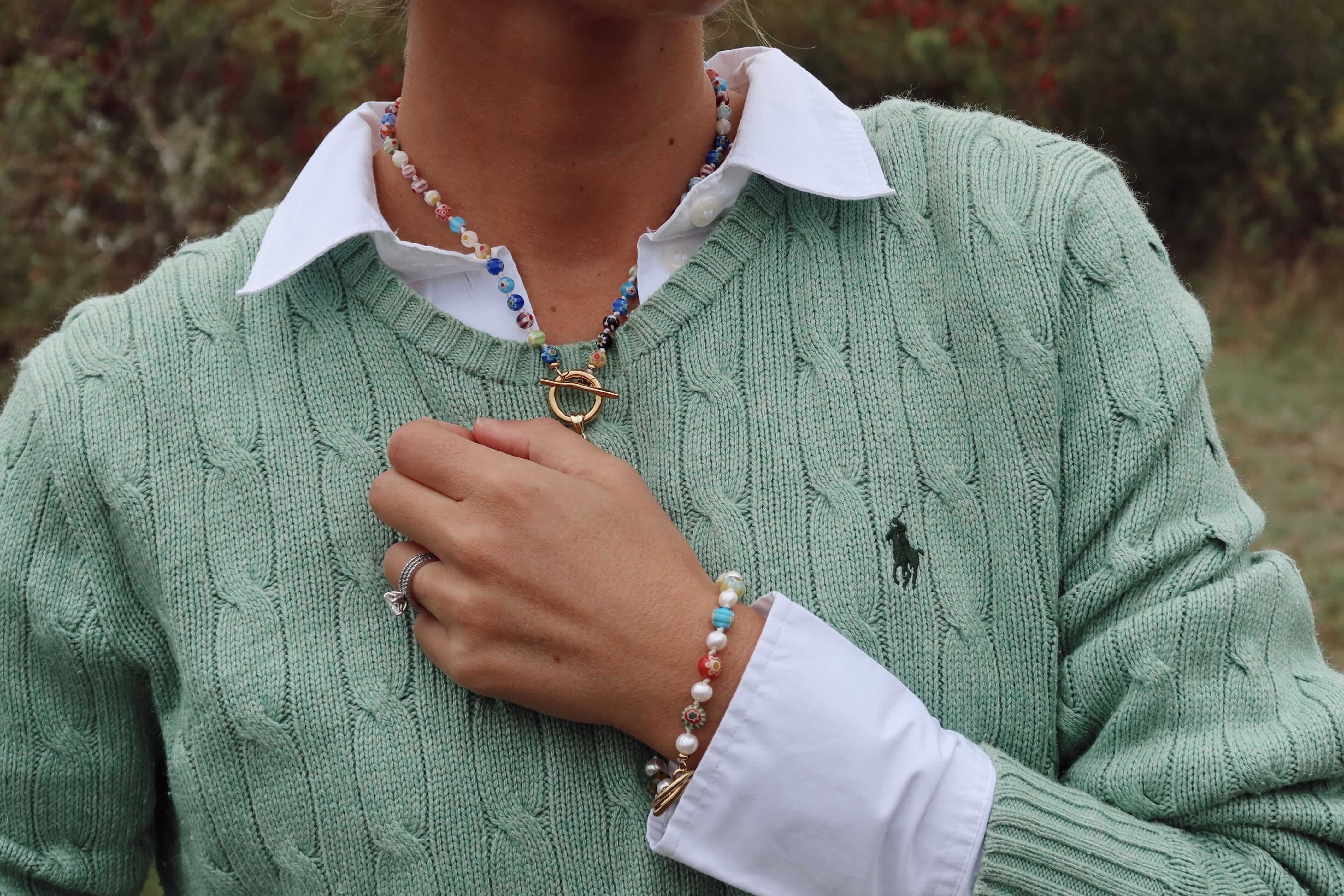A person wearing a sage green cable-knit sweater over a white collared shirt, with a beaded necklace and bracelet visible. The sweater.