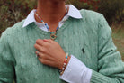 A person wearing a sage green cable-knit sweater over a white collared shirt, with a beaded necklace and bracelet visible. The sweater.