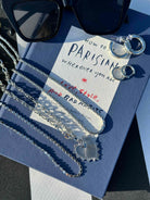 A styled flat-lay featuring a silver chain necklace with a heart-shaped pendant draped over a blue book. A white card with handwritten text lies on top of the book, alongside black sunglasses and silver key rings.