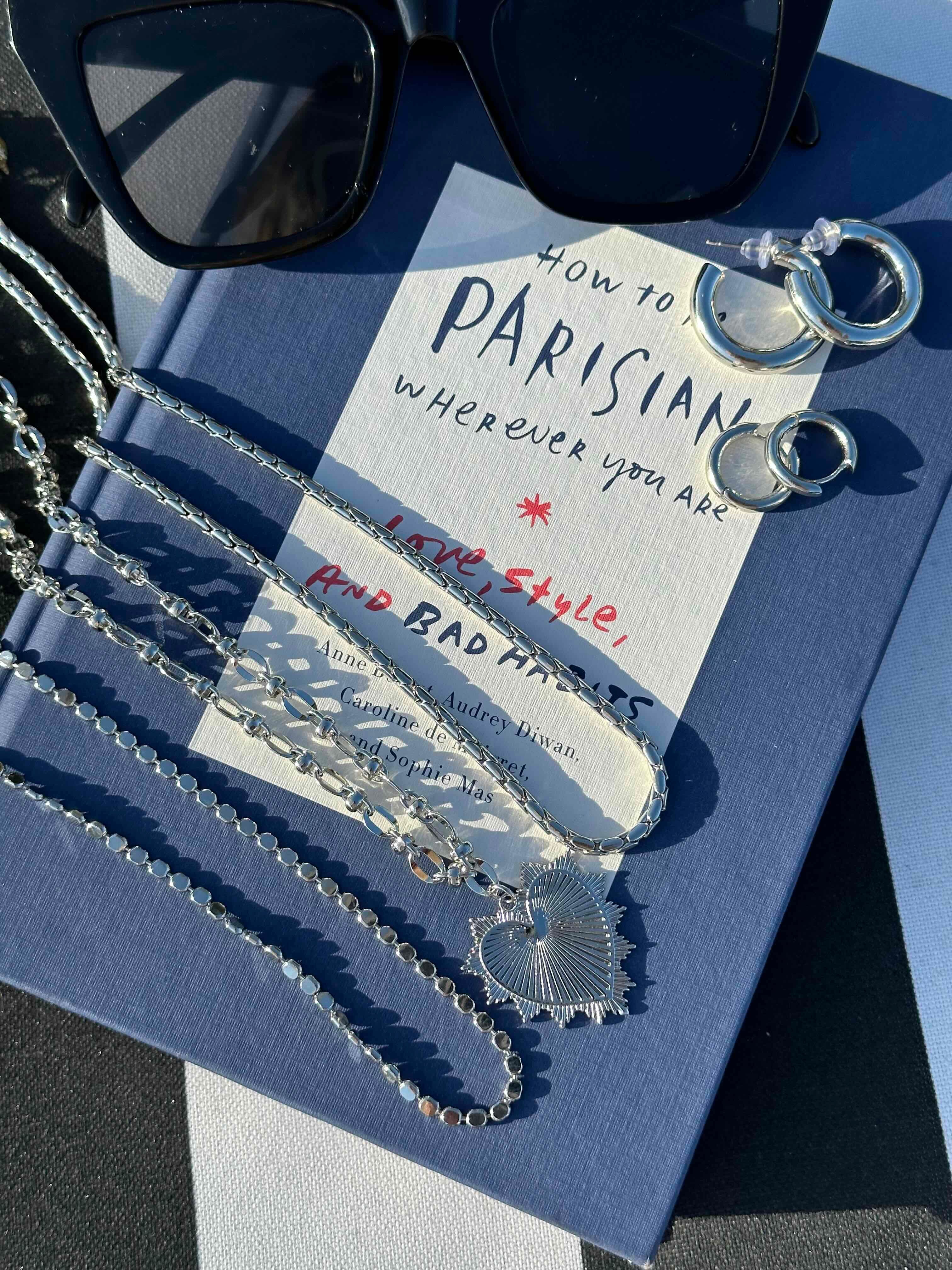 A styled flat-lay featuring a silver chain necklace with a heart-shaped pendant draped over a blue book. A white card with handwritten text lies on top of the book, alongside black sunglasses and silver key rings.