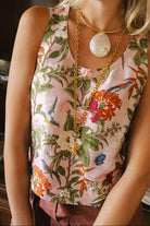 A woman wearing a sleeveless pink floral top with green leaves, orange flowers, and birds, accessorized with gold necklaces and sunglasses in a warm indoor setting.