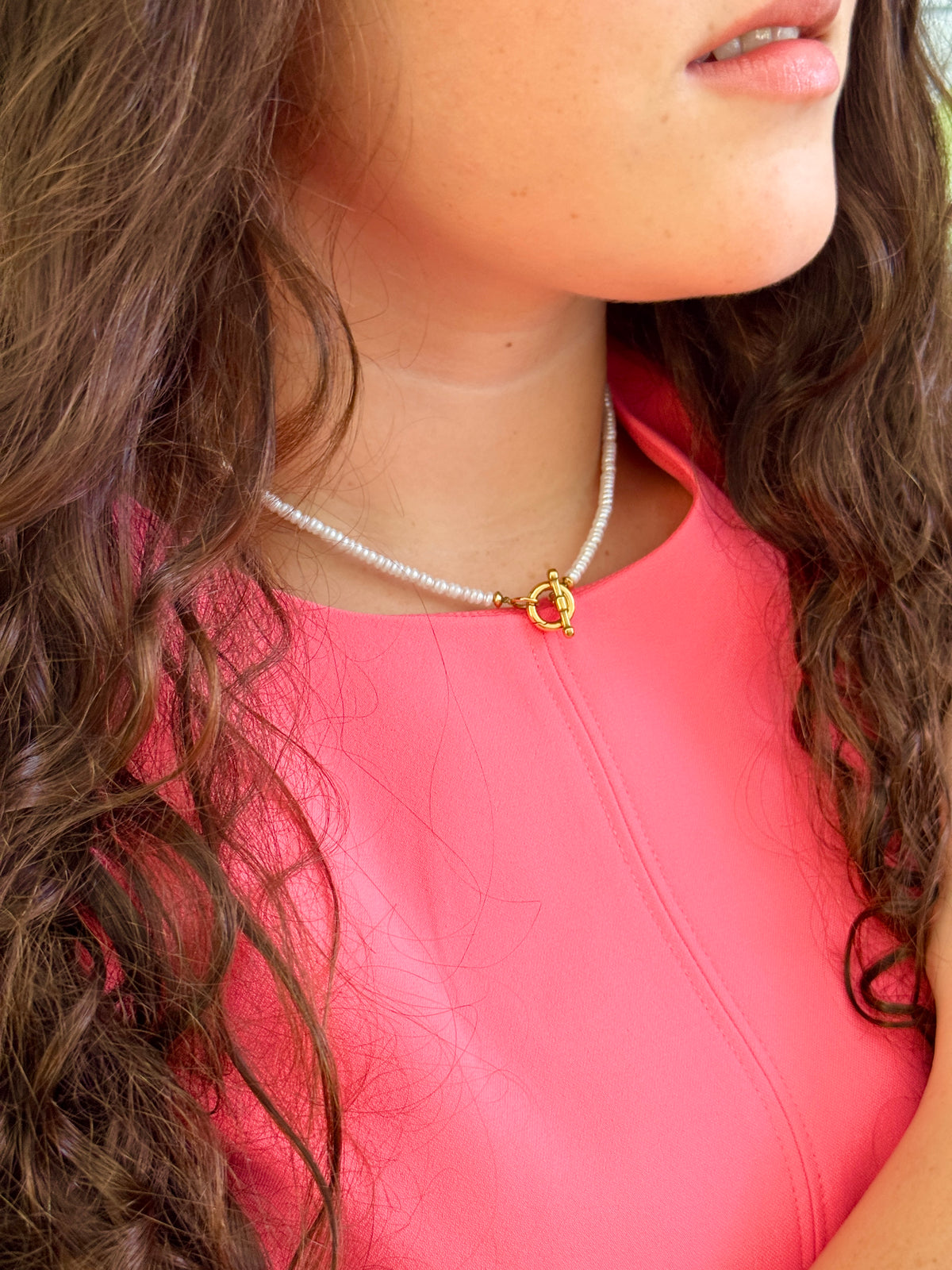 A close-up of a woman wearing a delicate white pearl necklace with a gold toggle clasp, paired with a bright pink top.