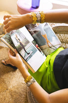 A close-up lifestyle shot of a person's arms and hands wearing two thick, decorative metallic bracelets while holding an open magazine. The bracelets feature a textured, layered design in gold and silver tones.