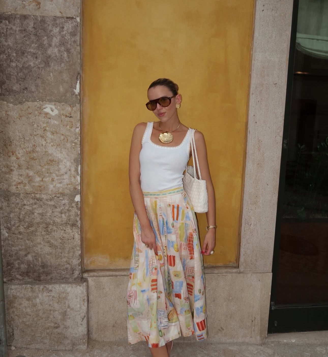 Woman wearing a white top and colorful skirt against a yellow wall.