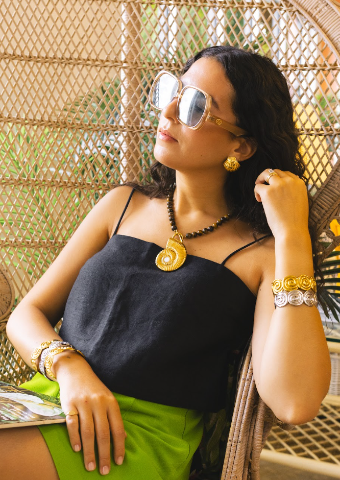 A woman wearing a black spaghetti-strap top and bright green skirt, sitting in a wicker hanging chair. She is wearing gold sunglasses, a gold spiral pendant necklace, and multiple gold and silver bracelets.