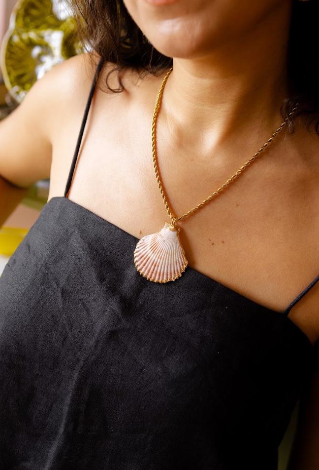 A close-up lifestyle shot of a woman wearing a gold chain necklace with a scallop shell pendant, paired with a black spaghetti strap top.