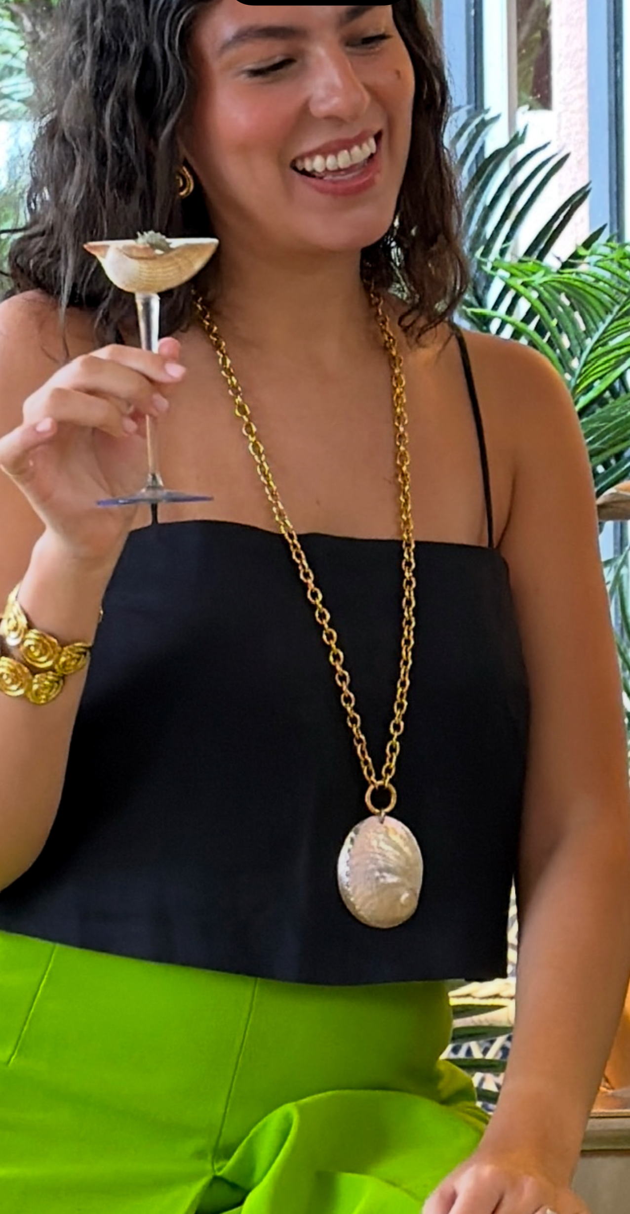 Woman holding a cocktail with a long gold necklace and green skirt.