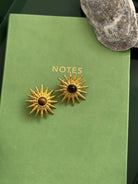 Gold sun-shaped earrings with a central brown gemstone on a green book.