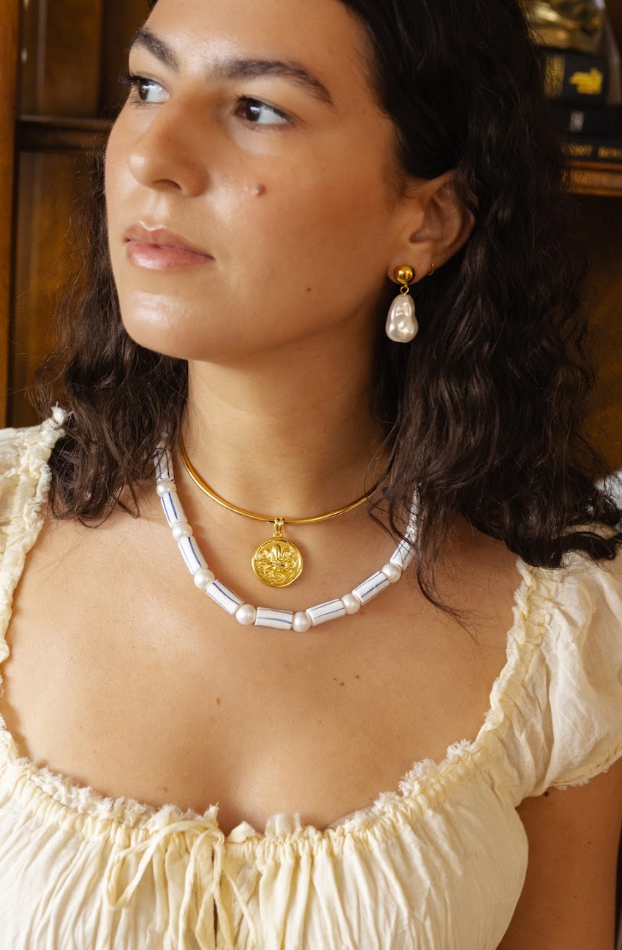 A woman wearing a gold necklace with a round medallion pendant and a white baroque pearl collar necklace, paired with a gold drop earring, in a warm indoor setting.