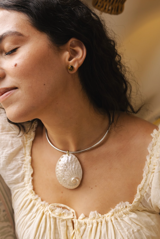 A close-up lifestyle shot of a woman wearing a silver-toned bangle necklace with a large, textured white seashell pendant. She is wearing a cream-colored ruffled blouse and a small gold stud earring.