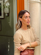 A woman wearing a beige knit crewneck sweater and a brown paperbag-style belt, standing next to a stone column.